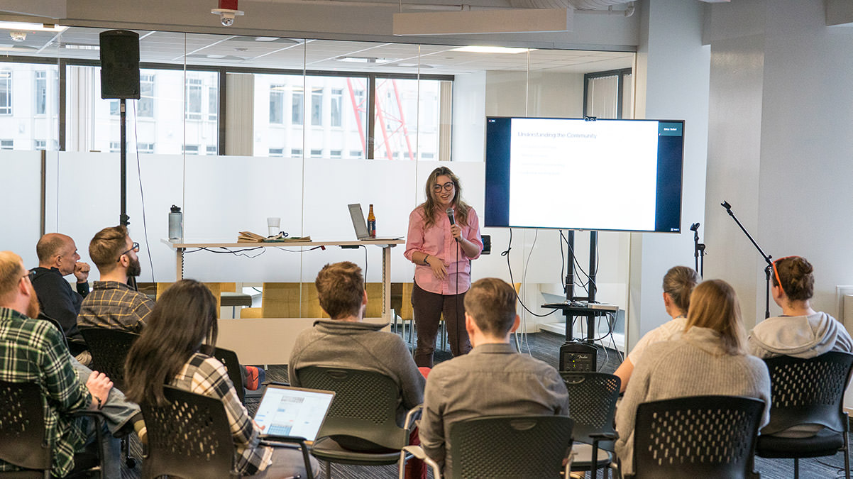 Woman presenting using large monitor and microphone in front of audience seated in moveable chairs in semicircle formation.