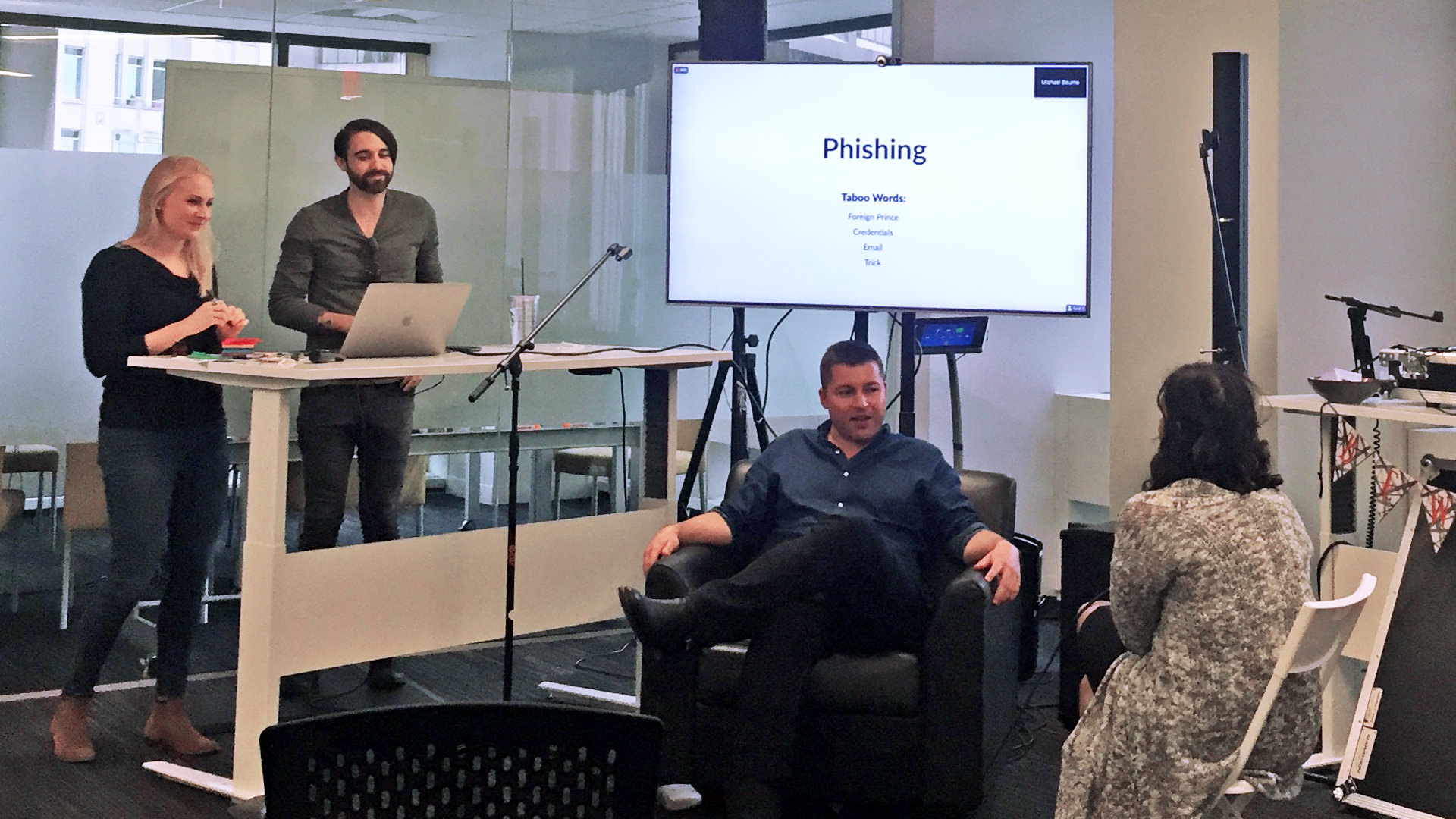 Two people run an activity in which two participants sit in chairs playing a data privacy themed version of the card game, taboo.